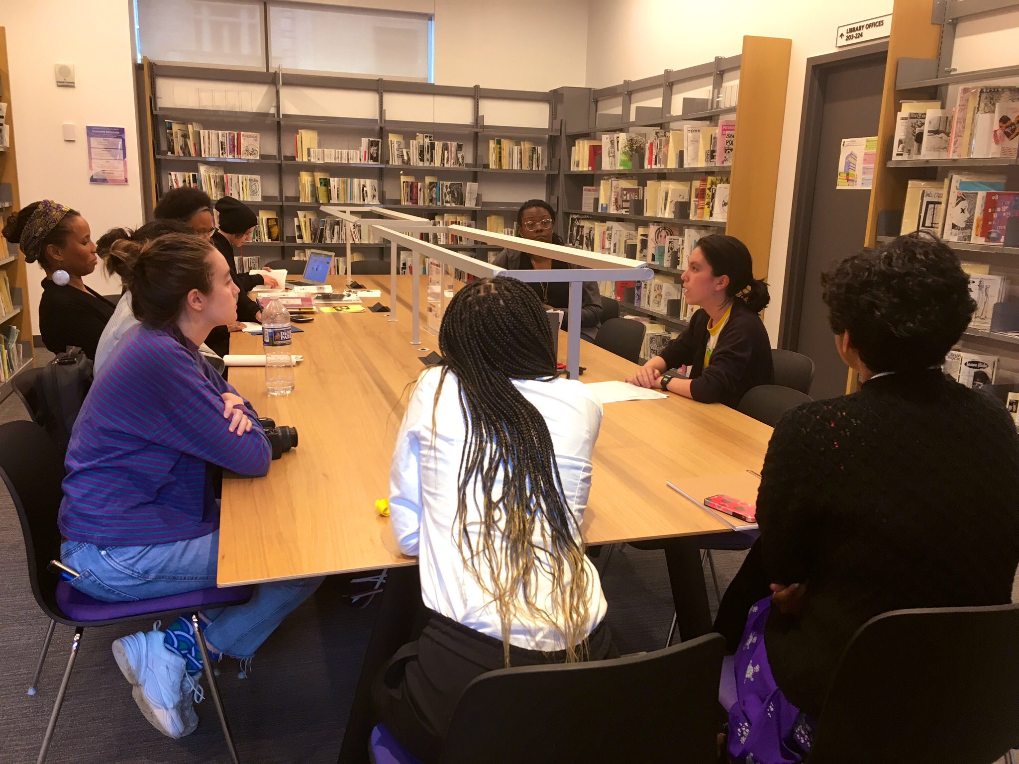 Summer Fellows sit around a long table in a room lined with bookshelves full of zines.