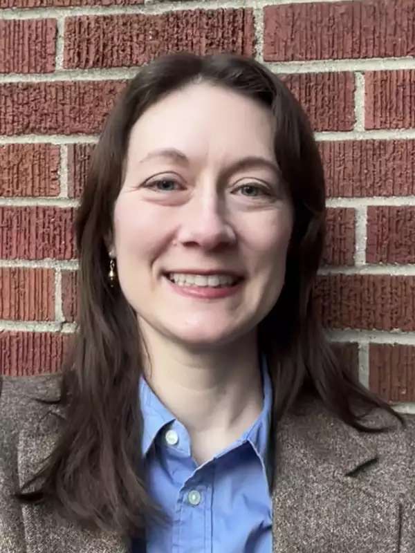 Profile photo of woman with brown hair in a tweed blazer in front of a brick wall.