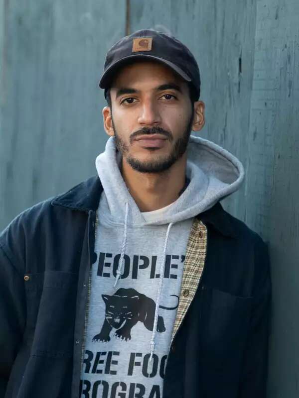 Photo of a man standing by an exterior wall, wearing a black hat and facing the camera. 